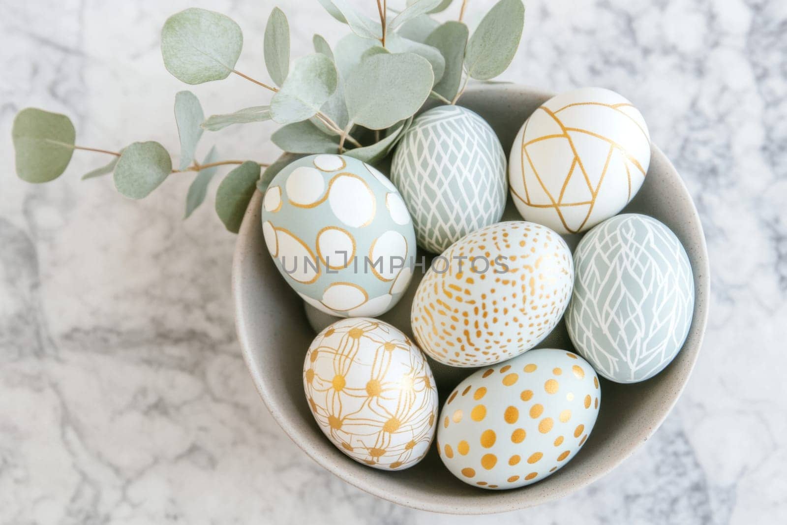 A stylish minimalist Easter centerpiece showcases beautifully decorated golden patterned Easter eggs arranged in a bowl. Eucalyptus leaves accent the display, set against a chic marble countertop.