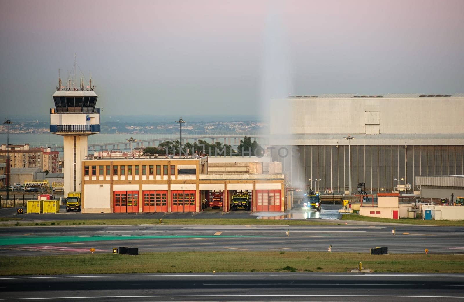 Lisbon, Portugal - May 9, 2024: Lisbon airport fire station with firefighting vehicles parked in front, next to ATC control tower and TAP Maintenance hangar. One vehicle doing water spray test, urban buildings at golden hour, safety and emergency