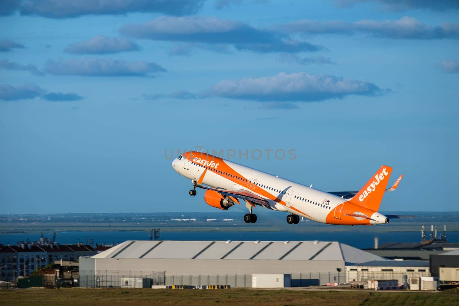 Lisbon, Portugal - Apr 29, 2024: Panning shot of EasyJet Airbus 321 neo aircraft take off from Lisbon airport runway. Cityscape, river view in background, airport warehouse facility, low-cost airline in clear blue sky and some clouds on sunny day