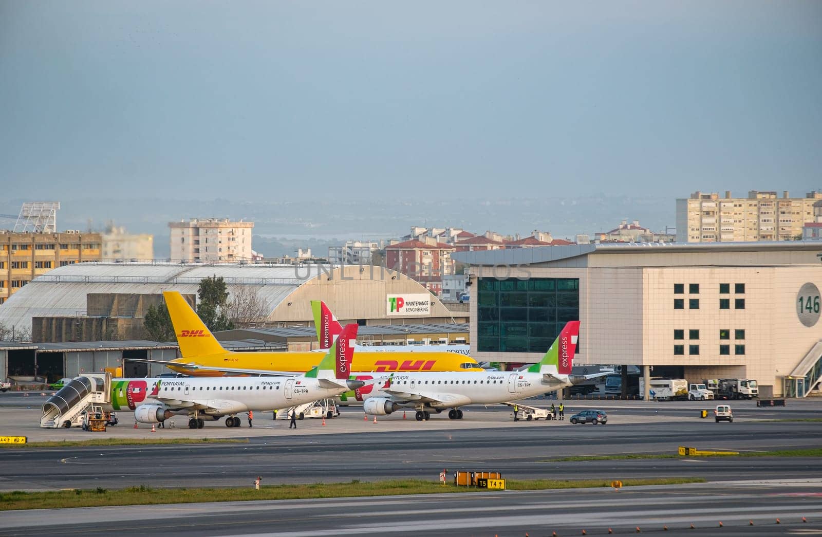 Lisbon, Portugal - Apr 3, 2024: TAP Express and DHL airplanes parked on apron at Lisbon Airport. TAP Maintenance and Engineering hangar, airport buildings and urban skyline in background on sunny day. Cargo and passenger aviation, busy modern airport