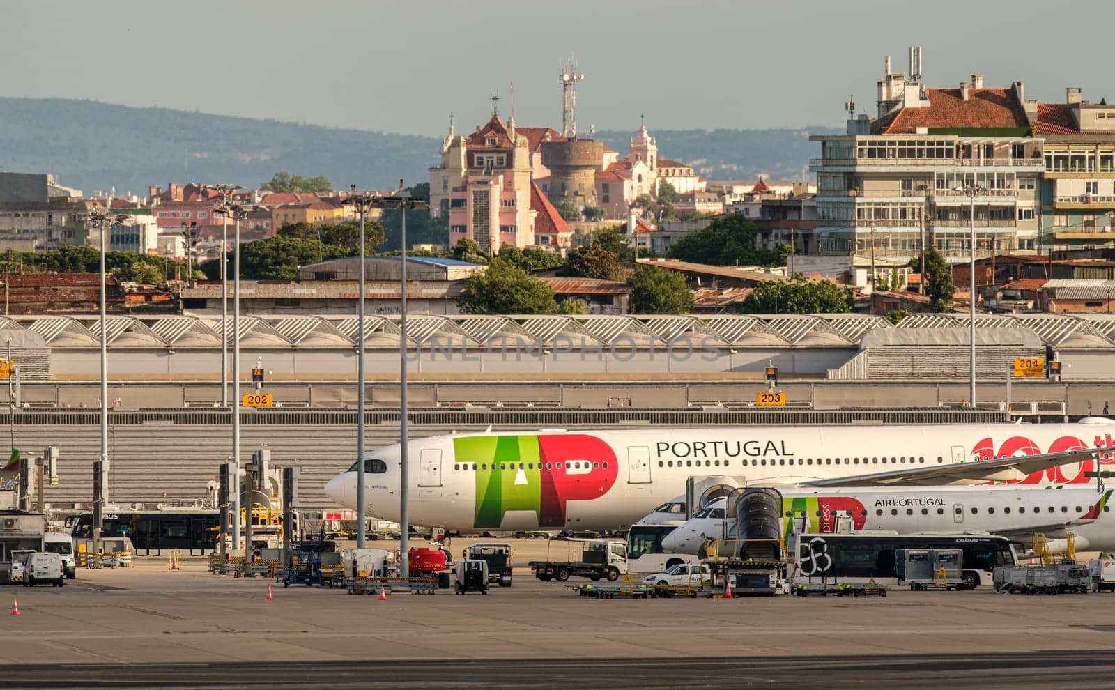 Lisbon, Portugal - Apr 24, 2024: TAP Air Portugal aircrafts waiting for boarding by airport hangars on sunny day. City landscapes in background, clear blue sky, summer haze, travel destination, vacations, transportation, Portuguese travel hub.