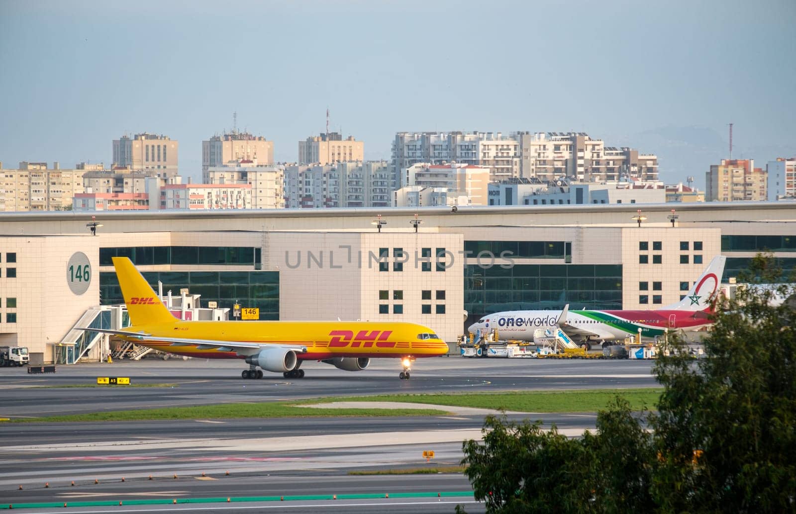 Lisbon, Portugal - Apr 3, 2024: DHL cargo airplane Boeing 757-200F taxiing at Lisbon Airport and Royal Air Maroc passenger airplane parked at nearby gate. Airport terminal, city skyline in background. International travel and shipment hub, sunny day