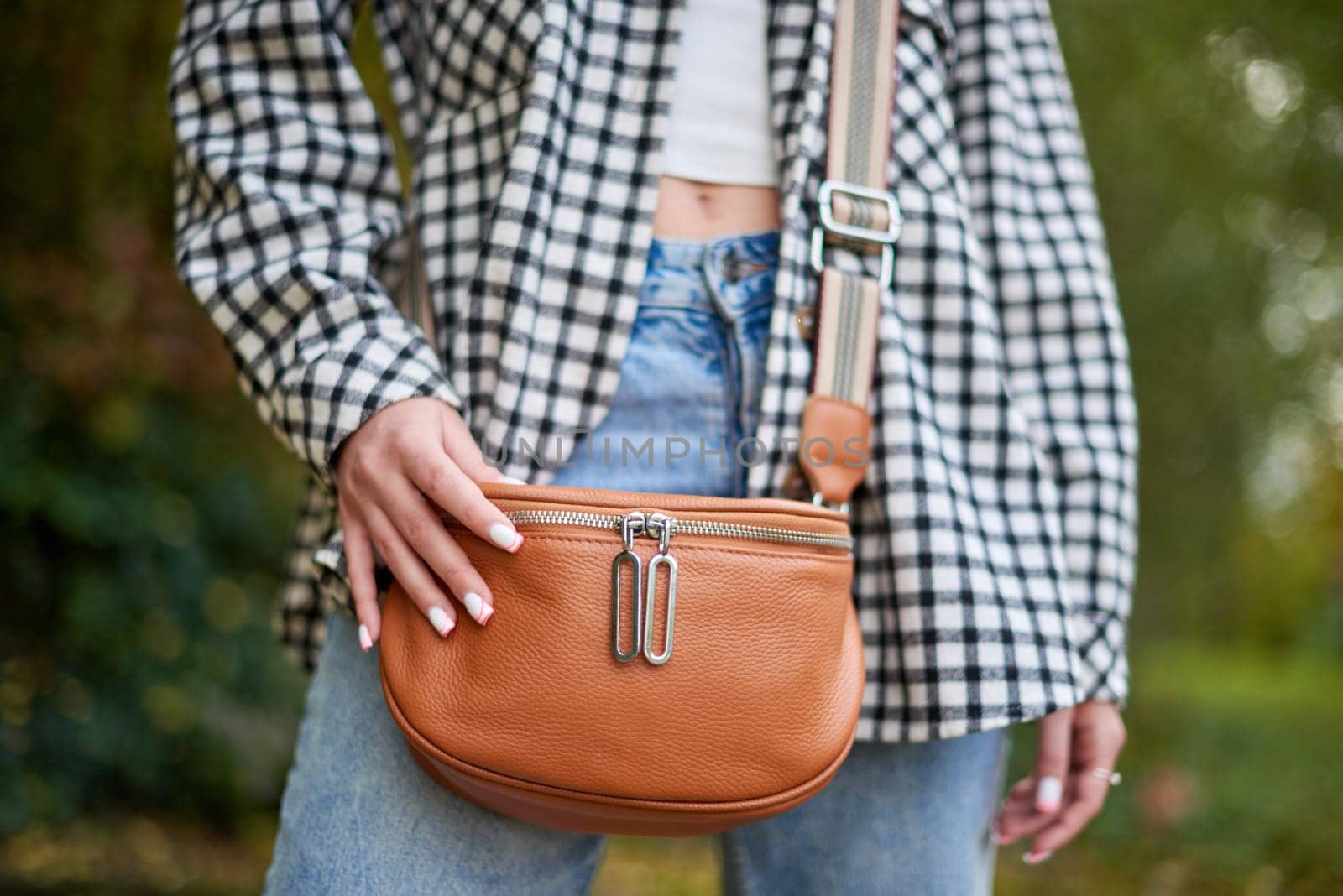 A young woman with wavy brown hair and blue eyes confidently poses outdoors in a lush autumn park. She wears a trendy black-and-white checkered shirt over a white crop top and classic blue jeans, accessorized with a tan leather crossbody bag with a striped strap. The soft natural lighting and blurred background of green foliage create a dreamy and cozy fall atmosphere. This fashionable portrait is ideal for lifestyle branding, autumn fashion campaigns, social media promotions, and advertising seasonal collections.