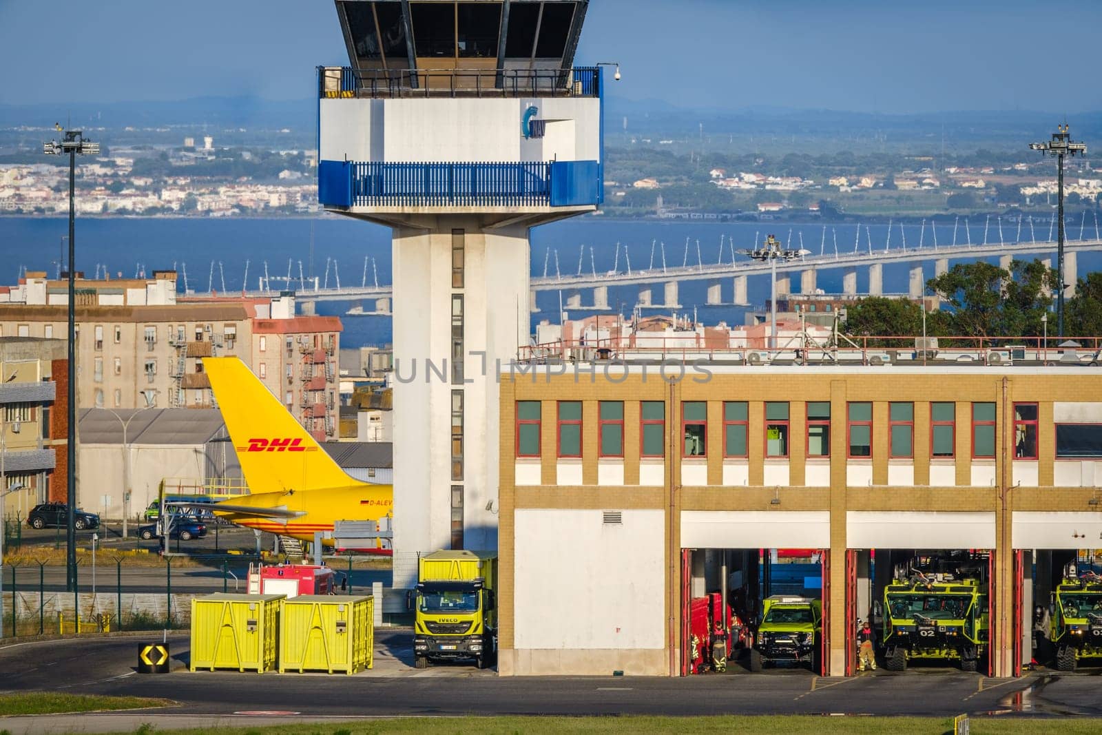 Lisbon, Portugal - Apr 24, 2024: Sunny day at Lisbon airport with tail of DHL cargo airplane, air traffic control tower and firefighters building in view. Clear blue sky, city and river in background. Ground vehicles, fire trucks ready for emergency.