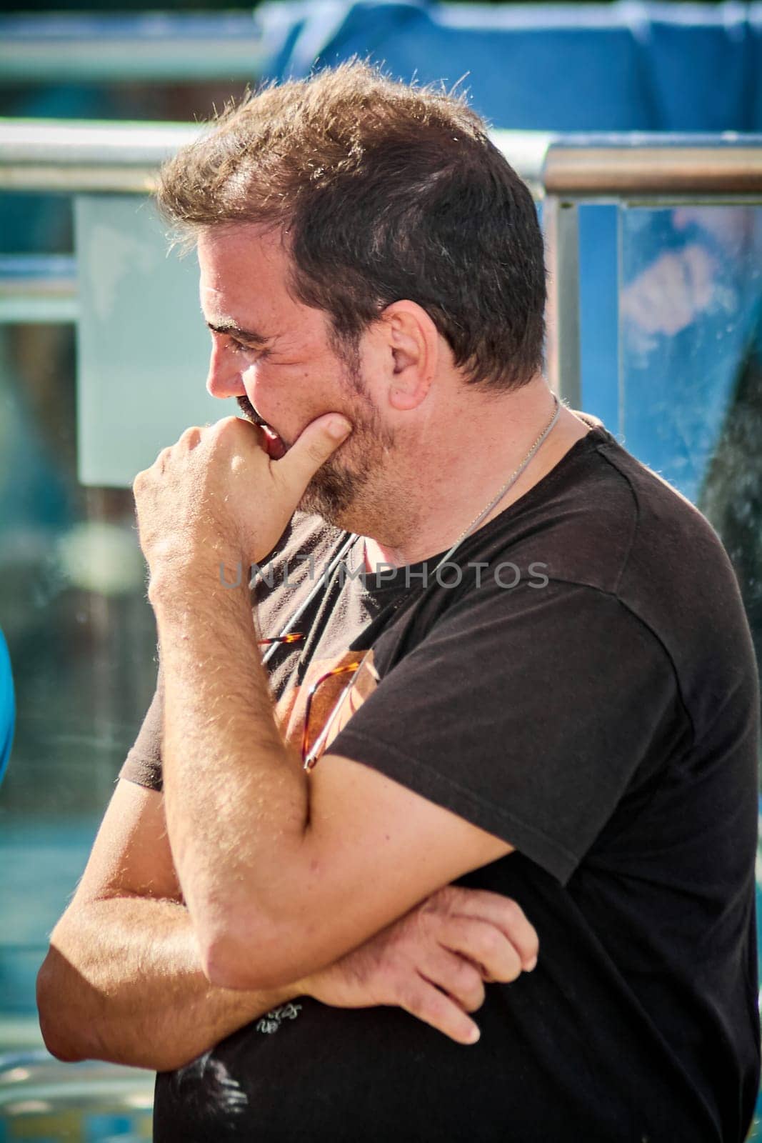A cheerful man wearing a black t-shirt hides his laugh behind his hand, framed by an urban outdoor environment with natural light.