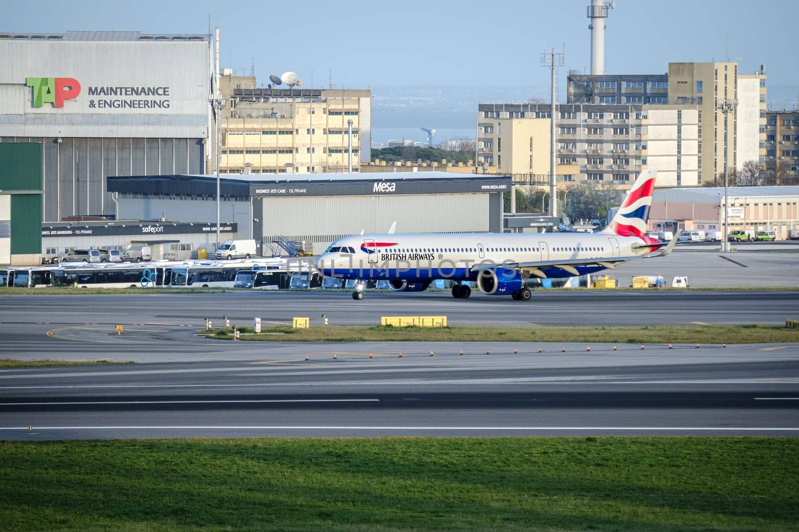 Lisbon, Portugal - Apr 3, 2024: British Airways airplane Airbus A321neo taxiing on runway at Lisbon Airport. TAP Maintenance and Engineering hangar, infrastructure, buses, city buildings in background. Clear blue sky, sunny day, travel destination