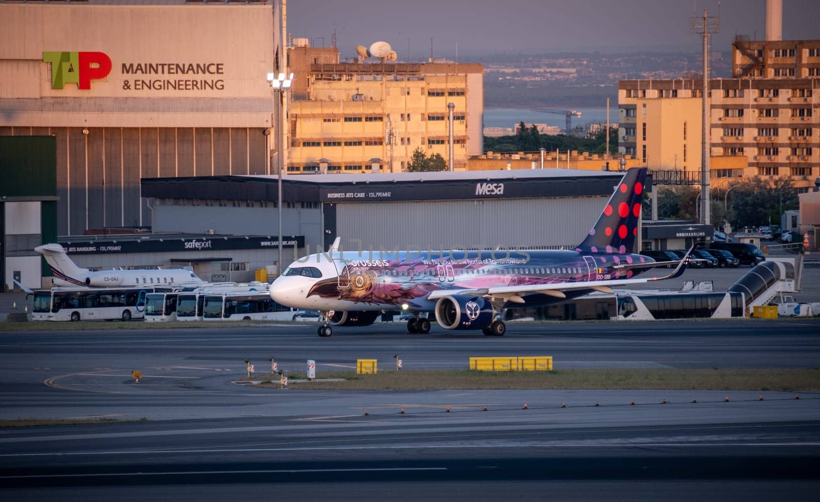 Lisbon, Portugal - May 9, 2024: Brussels Airlines Airbus A320 neo in colorful Amare livery inspired by Tomorrowland festival taxiing at Lisbon airport at golden hour. TAP Maintenance and Engineering hangar, buses, and urban buildings in background