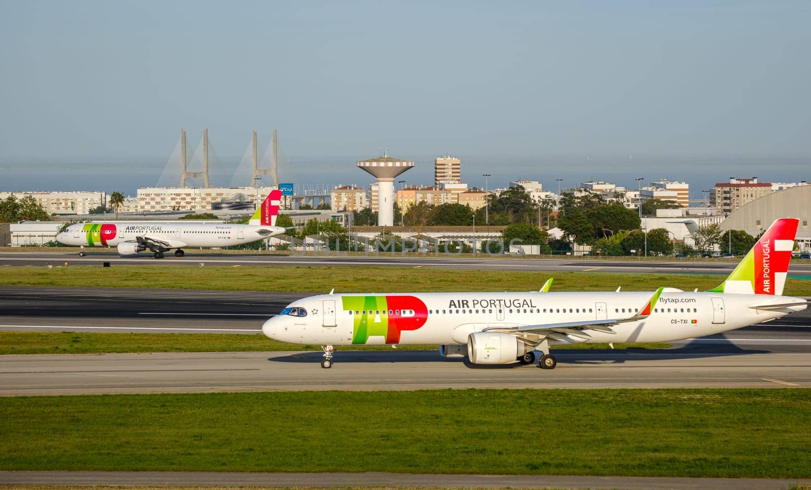 Lisbon, Portugal - Apr 3, 2024: Two TAP Air Portugal airline Airbus A321neo aircrafts ready for takeoff at Lisbon airport, clear blue sky, sunny day. Summer haze, vacation, travel destination, cityscape and Vasco da Gama bridge in background