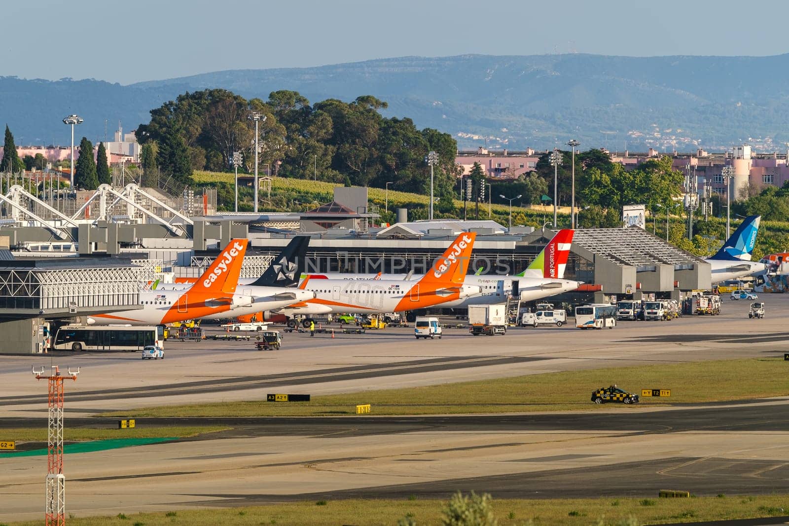 Lisbon, Portugal - Apr 24, 2024: TAP, Azores Airlines and EasyJet airplanes in Lisbon airport ready for boarding and takeoff on sunny day, clear blue sky. Ground support vehicles, airport Terminal 1 infrastructure, city buildings in background.