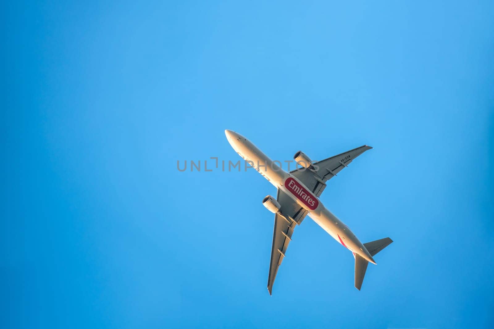 Lisbon, Portugal - Apr 3, 2024: Upshot of Emirates Boeing 777-300ER airplane flying overhead against clear blue sky on sunny day. Aircraft, airline, branding or logo, bottom fuselage, sunlight. Passenger long-haul flight, flag carrier from Dubai, UAE