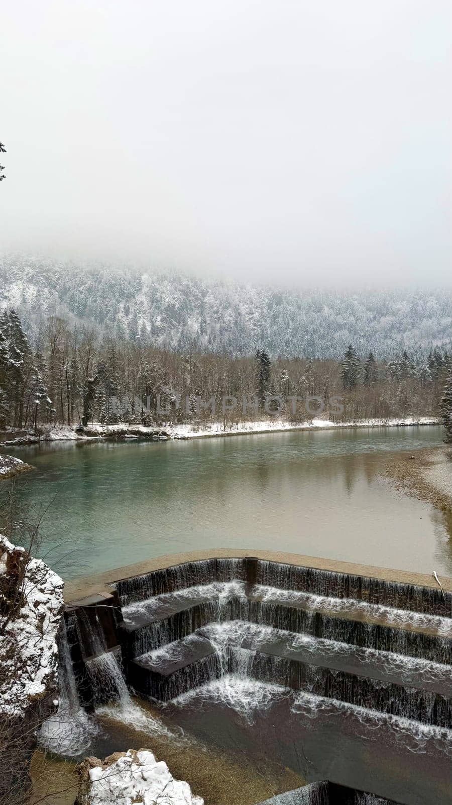 Tiered Waterfall Lechfall in a Snowy Mountain Landscape in Fuessen, Germany. A beautiful multi-tiered waterfall cascading over dark stone steps into a turquoise river Lech, surrounded by a snowy forest. The misty Alpine mountain in the background adds a mystical atmosphere to the winter scenery.