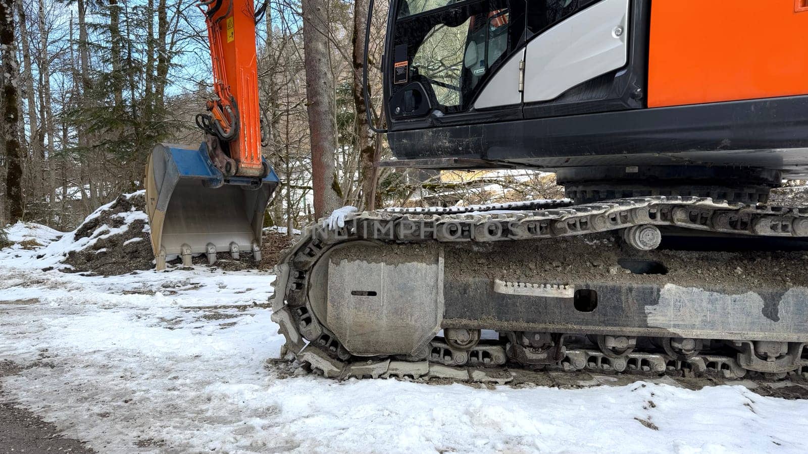 Orange Excavator on Snowy Ground With Tracks and Mechanical Bucket in Forest. An orange excavator with caterpillar tracks and a mechanical bucket is on snowy terrain surrounded by trees. Perfect for themes of construction, heavy machinery, winter work, or industrial operations.