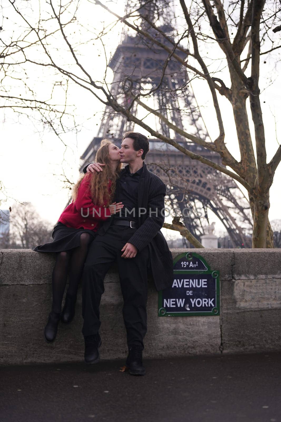 A young couple enjoys a romantic moment on Avenue de New York in Paris, with the Eiffel Tower towering behind them. The woman leans in for a kiss, capturing an intimate and stylish scene.