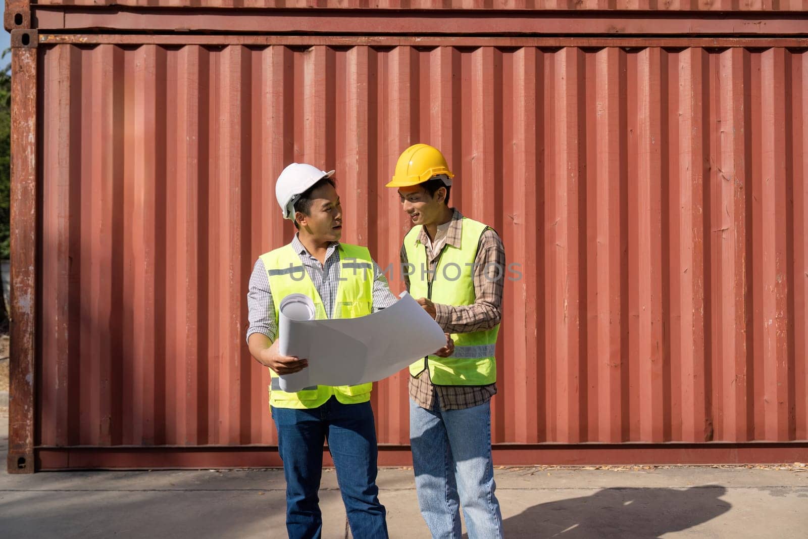 Two engineers in safety gear are reviewing plans and discussing logistics in a construction site environment, showcasing teamwork and collaboration.