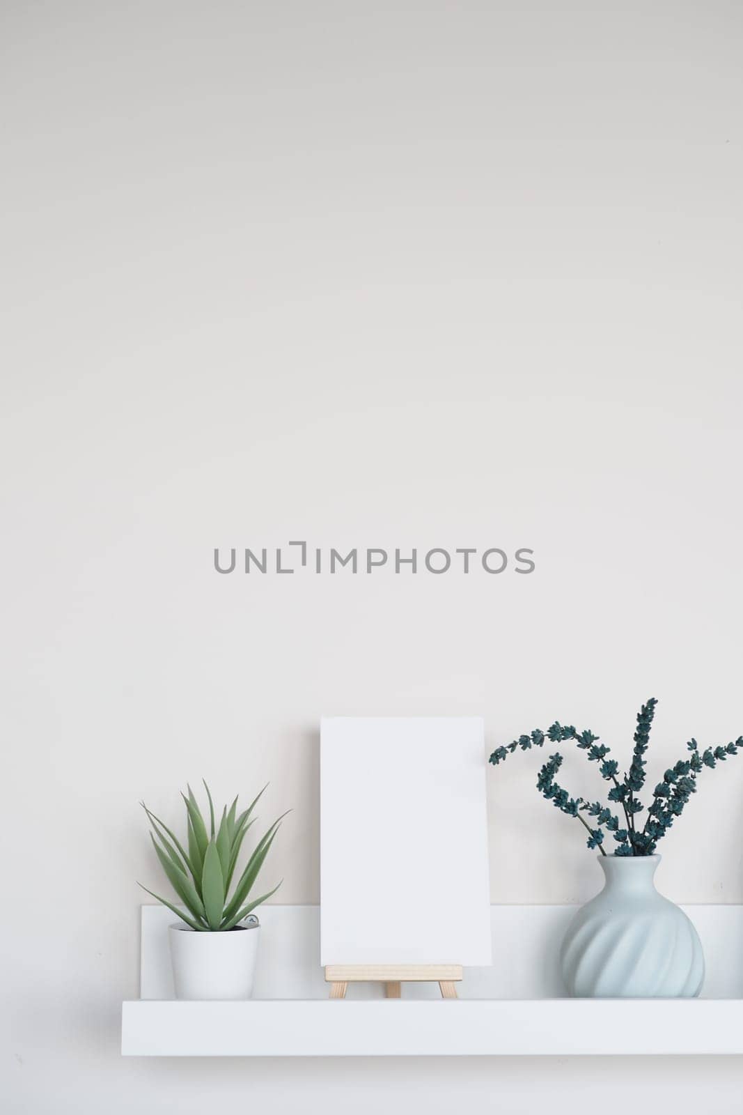 A clean shelf holds a blank canvas, green plants, and a vase.