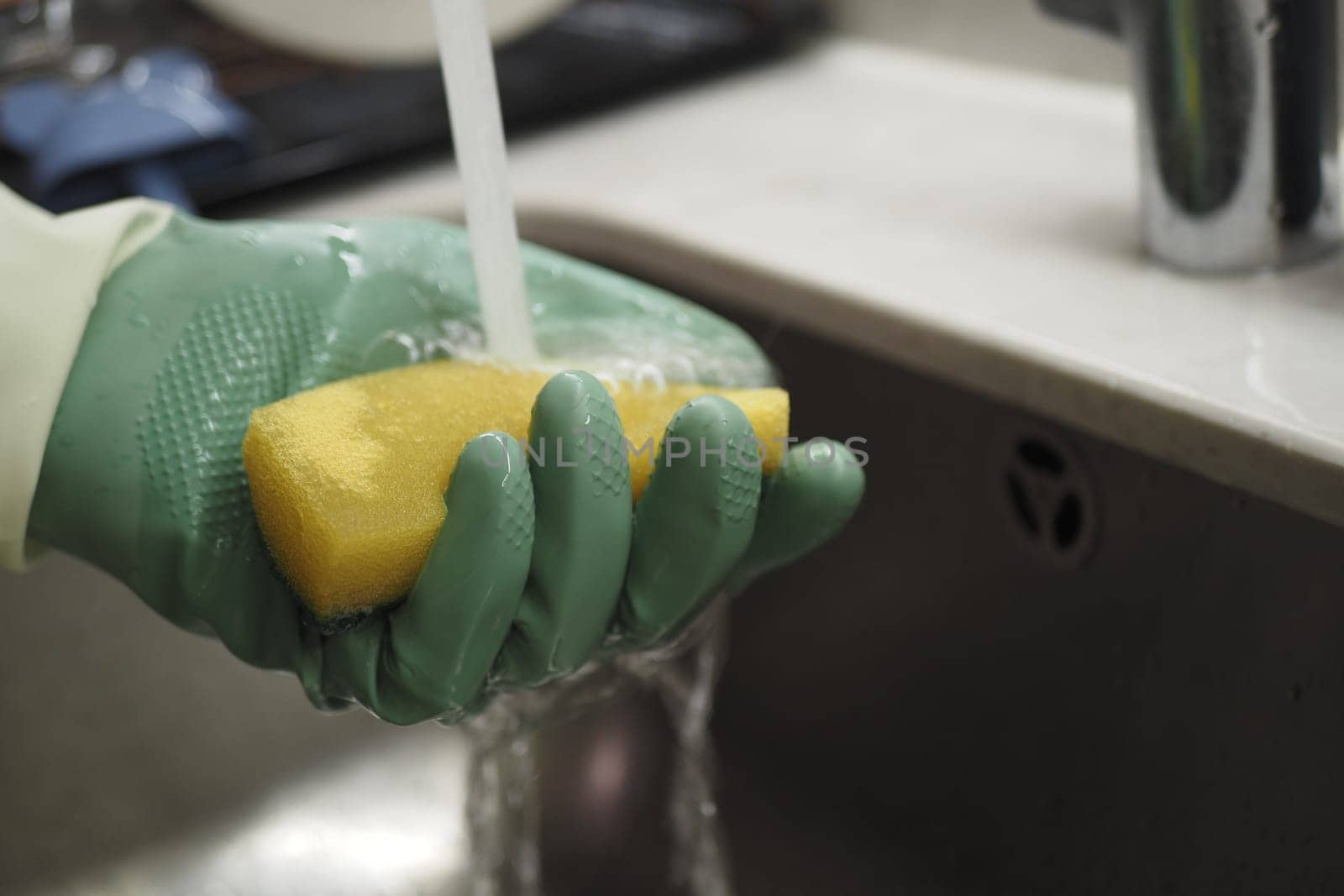 A person rinses a yellow sponge while wearing green gloves.