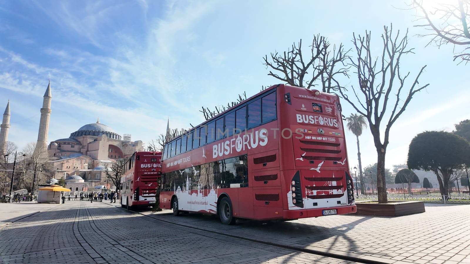 Tourists board red sightseeing buses next to Hagia Sophia under clear skies.