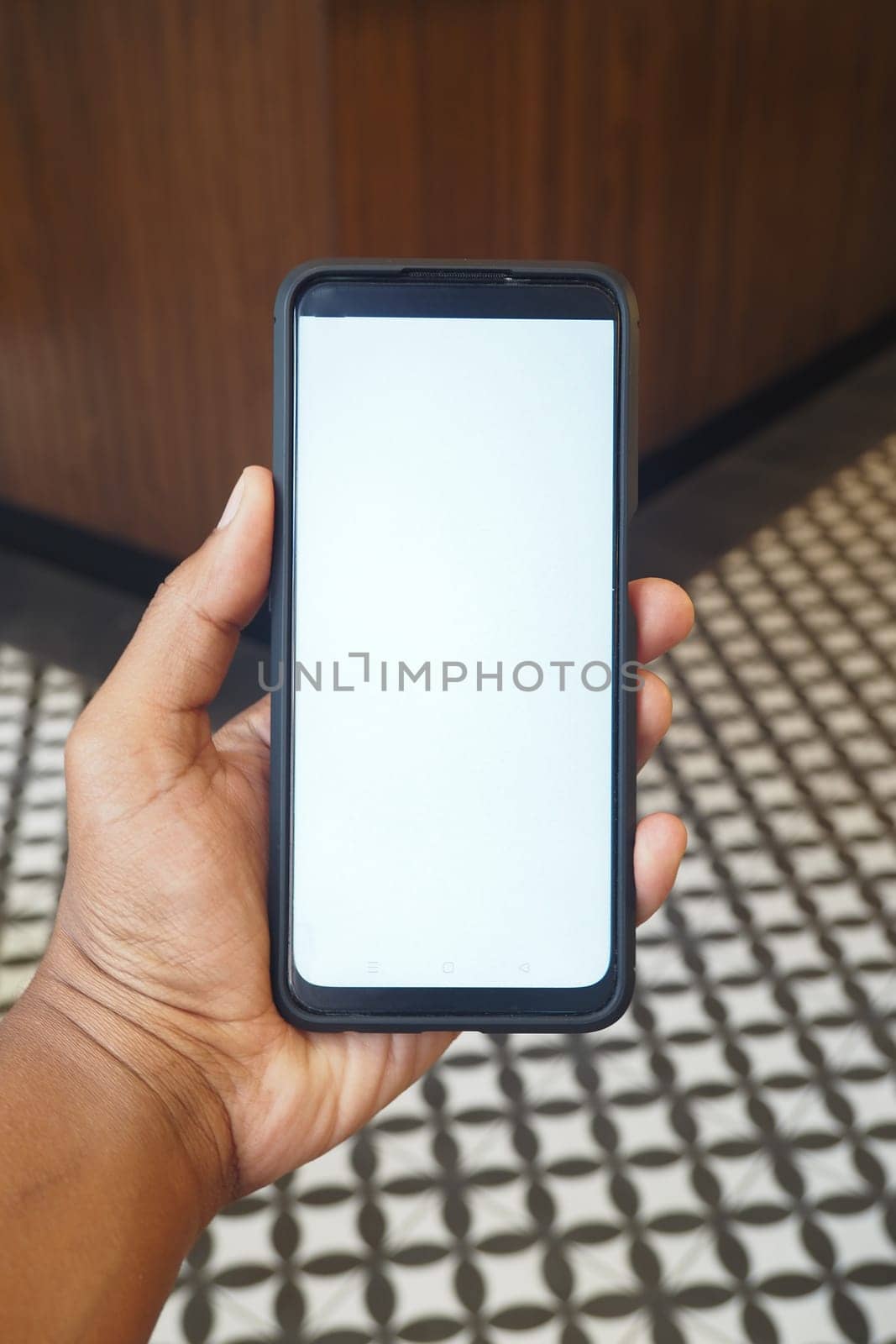 A person is holding a blank smartphone in a trendy cafe with patterned tiles.