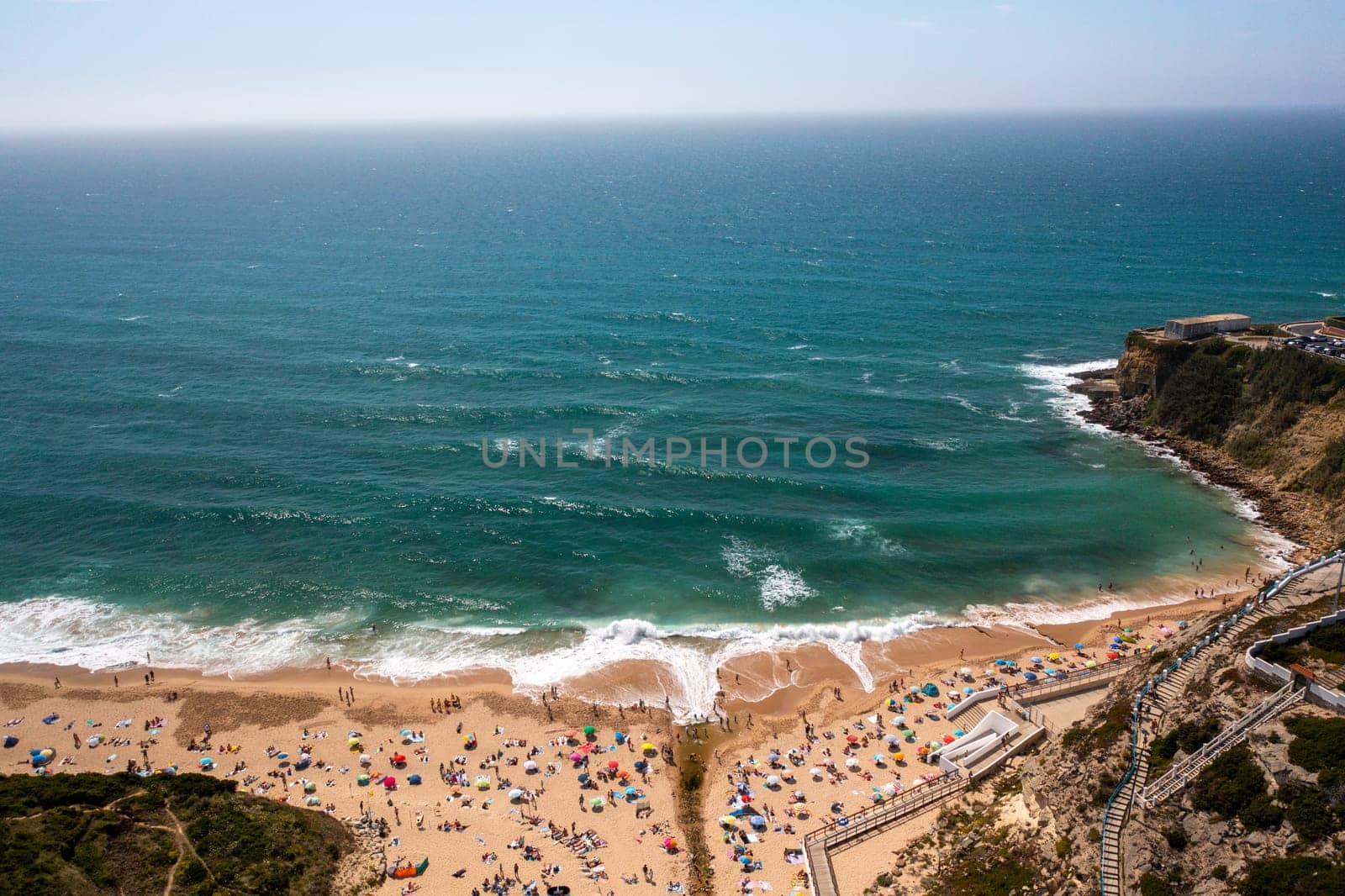 Aerial drone view of tourists on sandy seashore at Magoito Beach. Sea waves splash on the coast of a famous destination in Sintra, Portugal, during summer