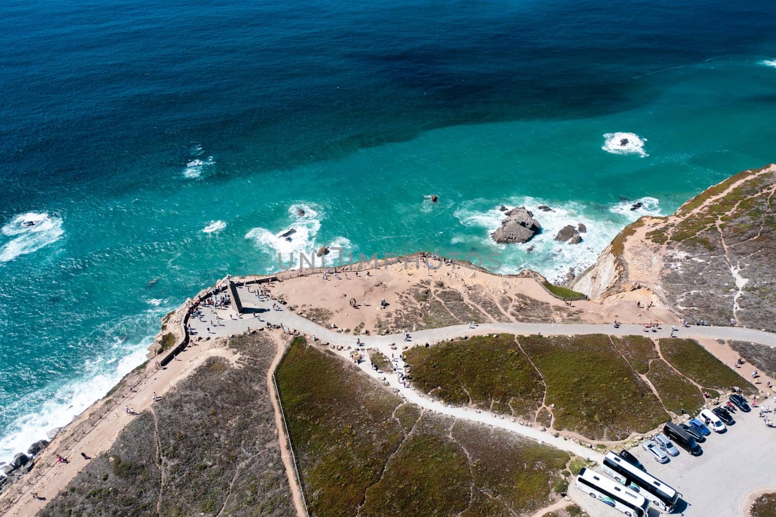 Aerial drone view of tourists exploring Cabo da Roca monument with stunning ocean waves and rocky cliffs, showcasing Portugal's scenic coastline