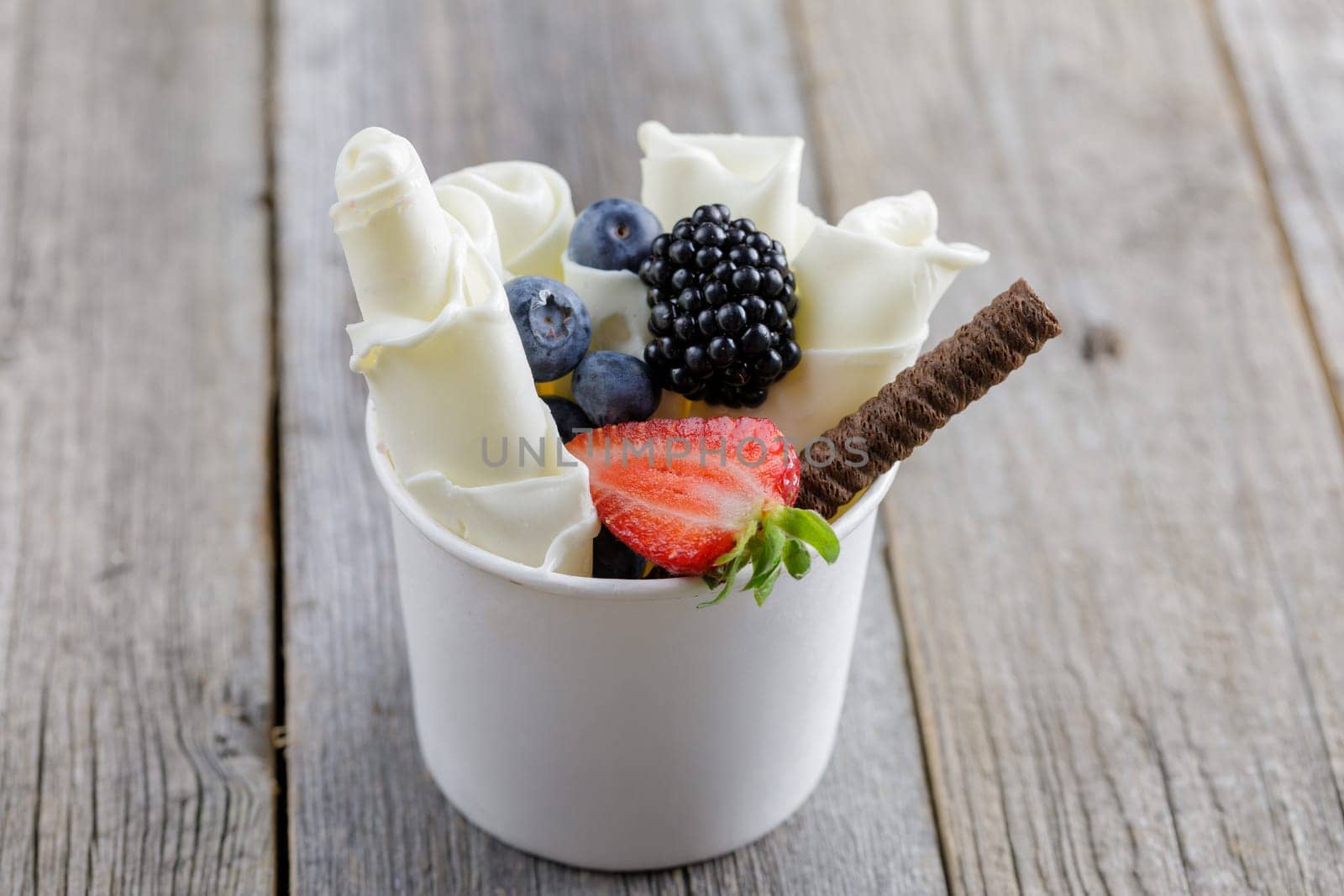 Delicious rolled ice cream in a cup, garnished with fresh berries, a chocolate wafer stick, and fruit toppings, served on a rustic wooden table