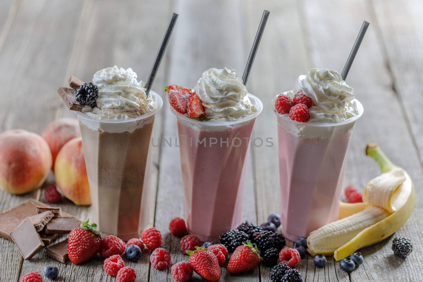 Three delicious milkshakes with whipped cream, fresh berries, chocolate, and fruit, served in tall cups on a rustic wooden table with ingredients.