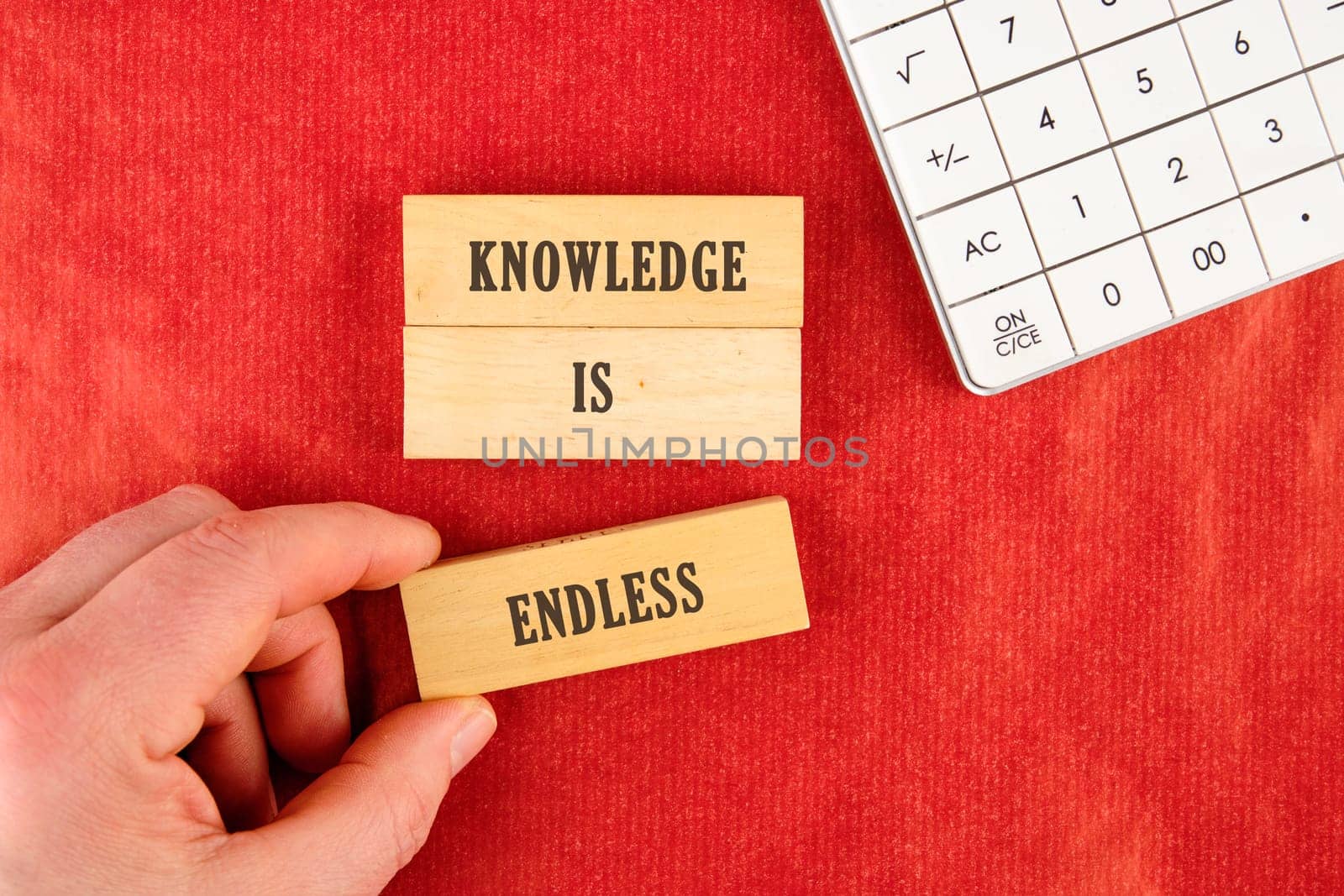 Hand is positioning wooden blocks that spell knowledge is endless on vibrant red surface next to a simple calculator