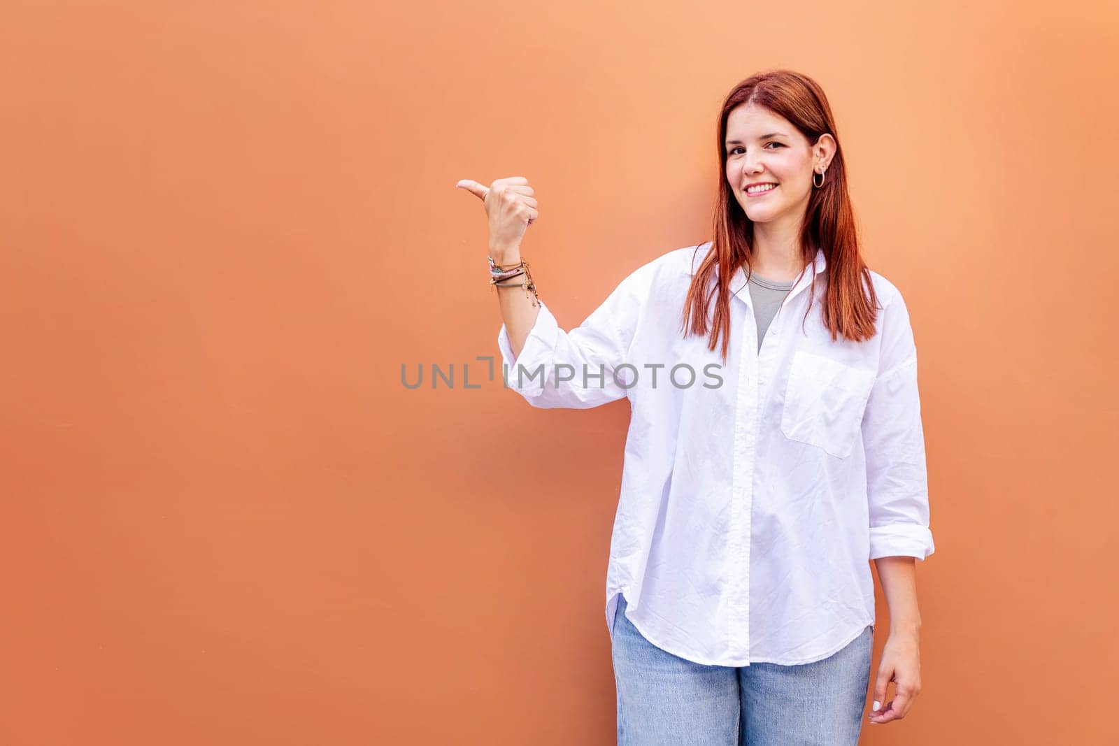 smiling young woman in white shirt and blue jeans is pointing to the side with the thumb while looking at camera, copyspace for text