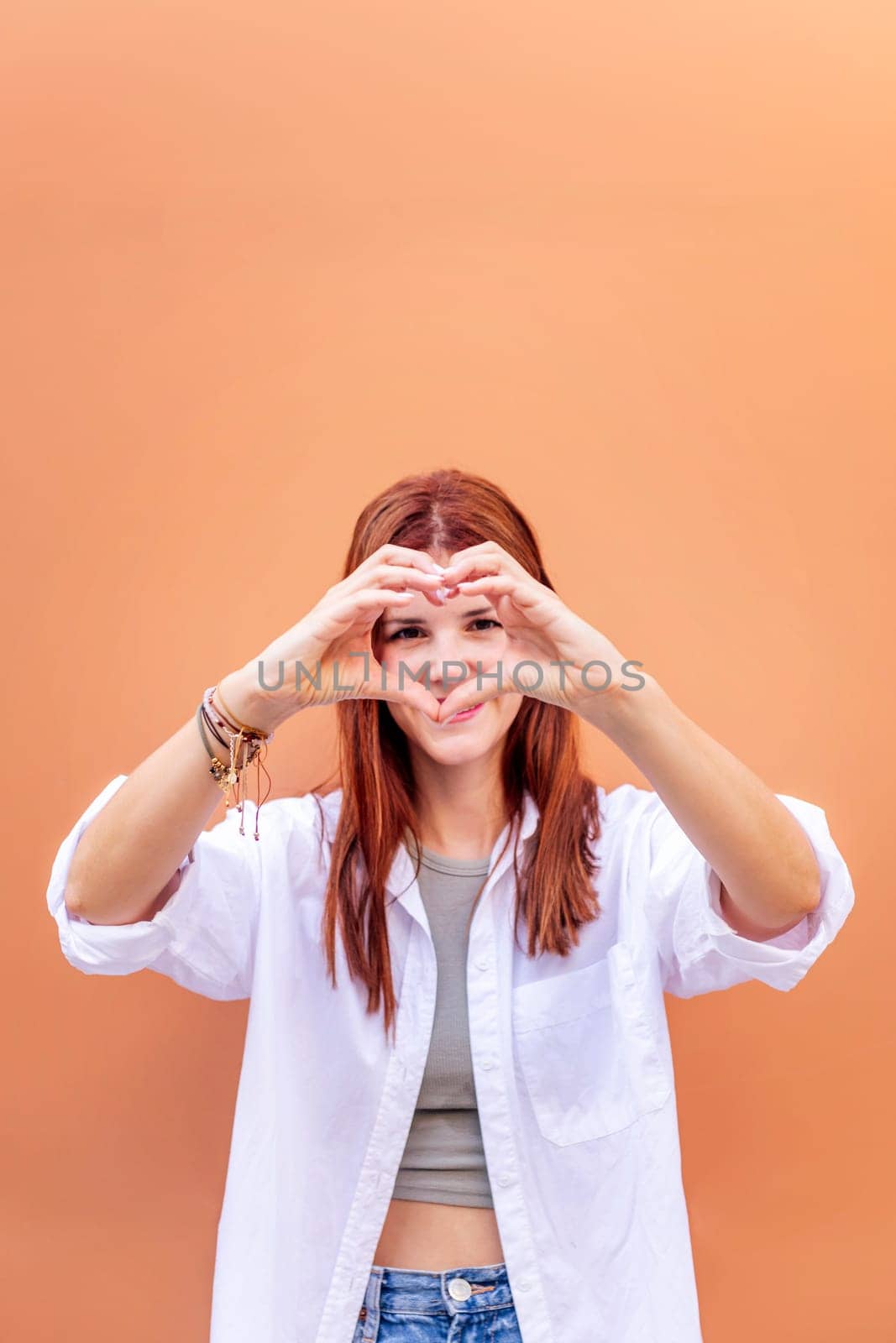 young woman standing in front of orange wall doing a heart shape with hands while looking at camera, she is wearing a white shirt, concept of love, copyspace for text
