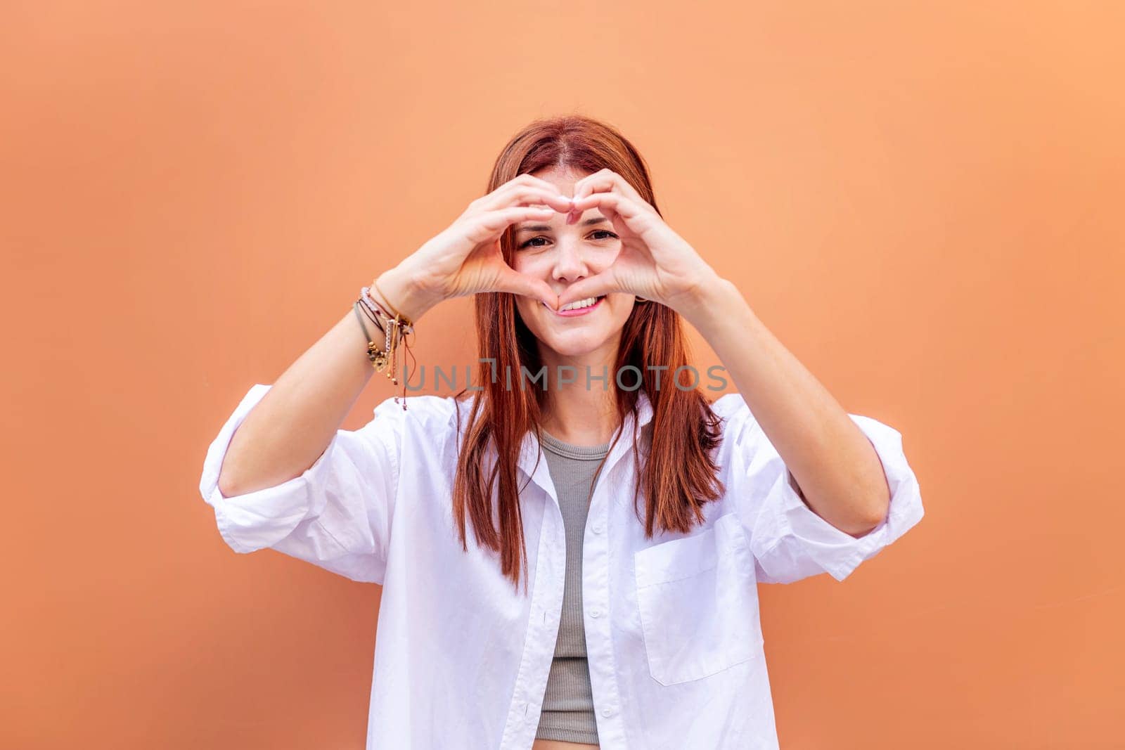young woman standing in front of orange wall doing a heart shape with hands while looking at camera, she is wearing a white shirt, concept of love, copyspace for text