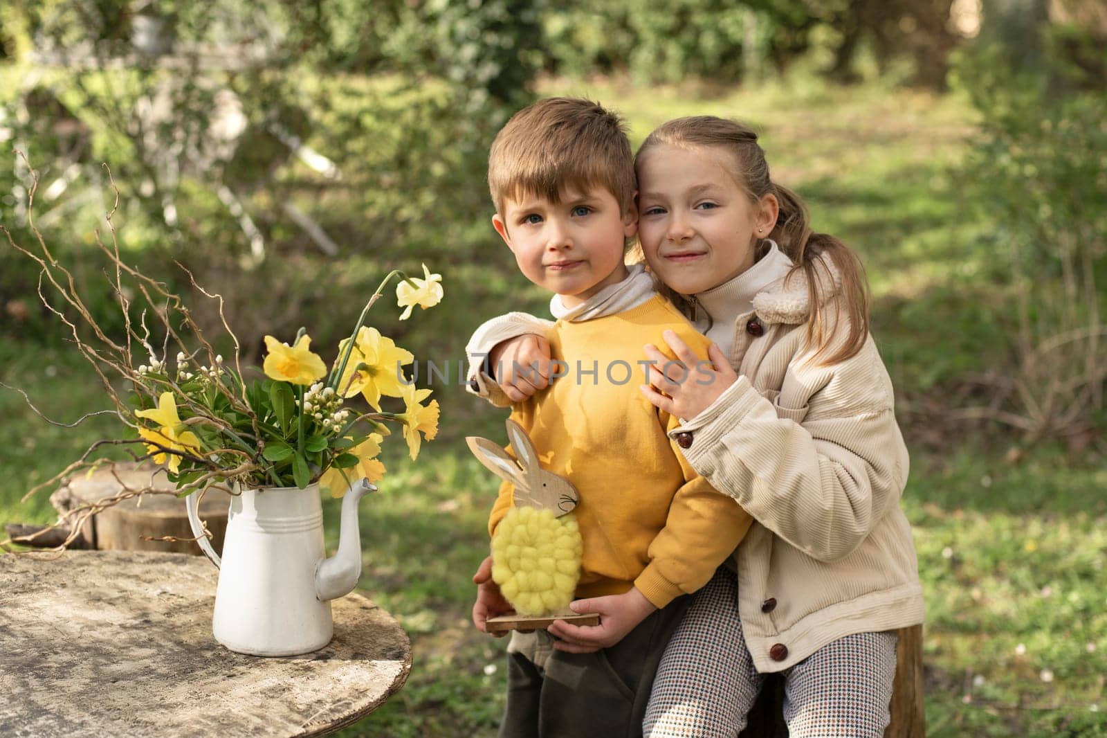 The children in a garden holds a yellow woolen rabbit. On a wooden table is a vase with daffodils. Easter