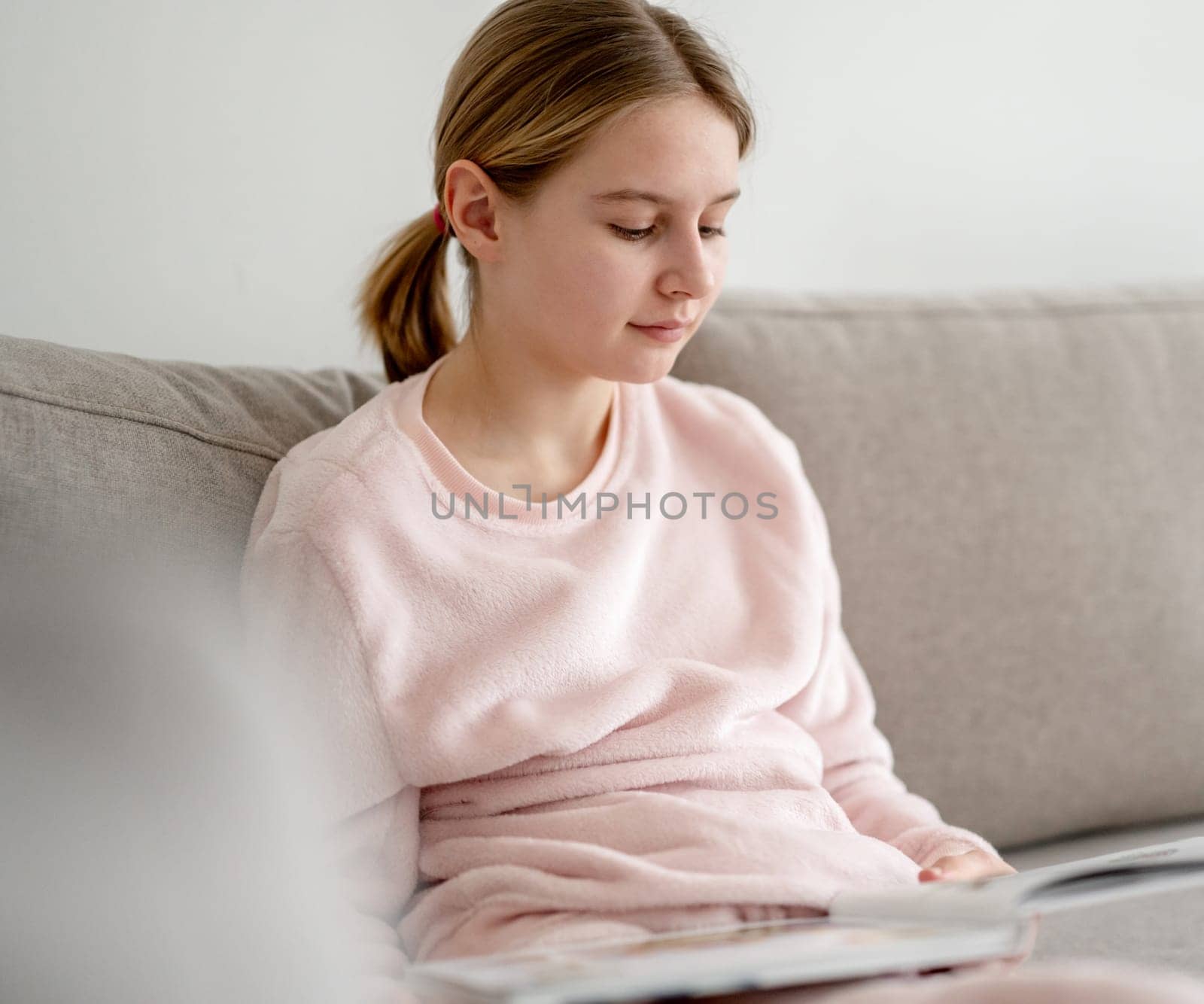 Little Girl Reads A Book At Home On The Sofa