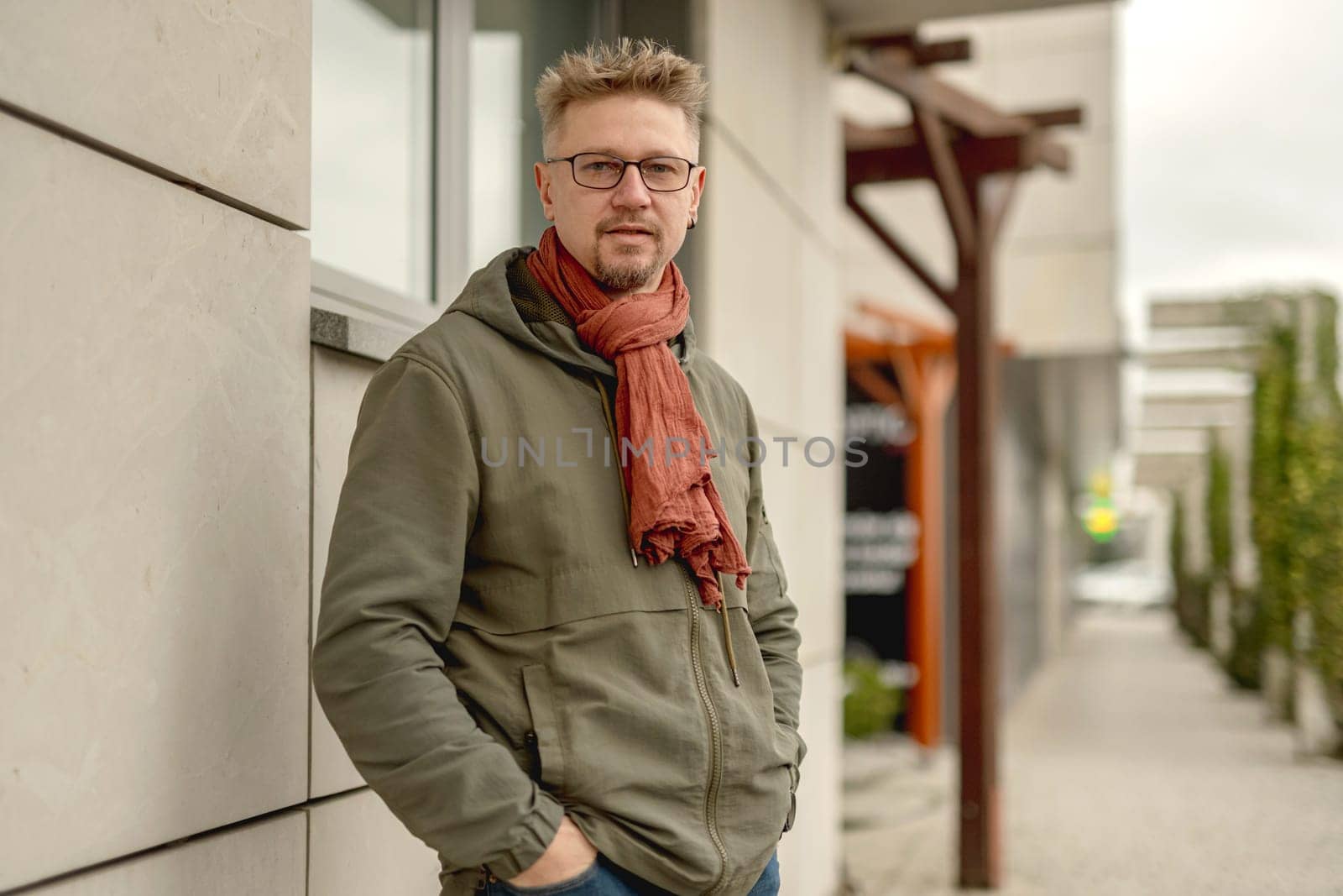 Young Attractive Man On A Street Wears Glasses And A Scarf, Looking Into The Camera