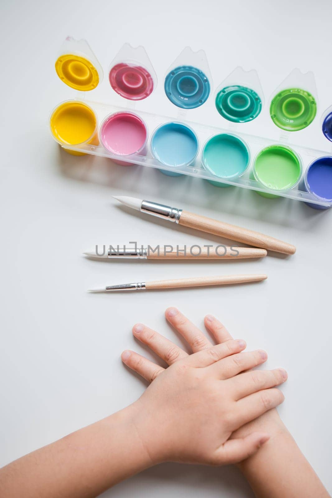 Childs hands with watercolor paints and brushes on a white surface.
