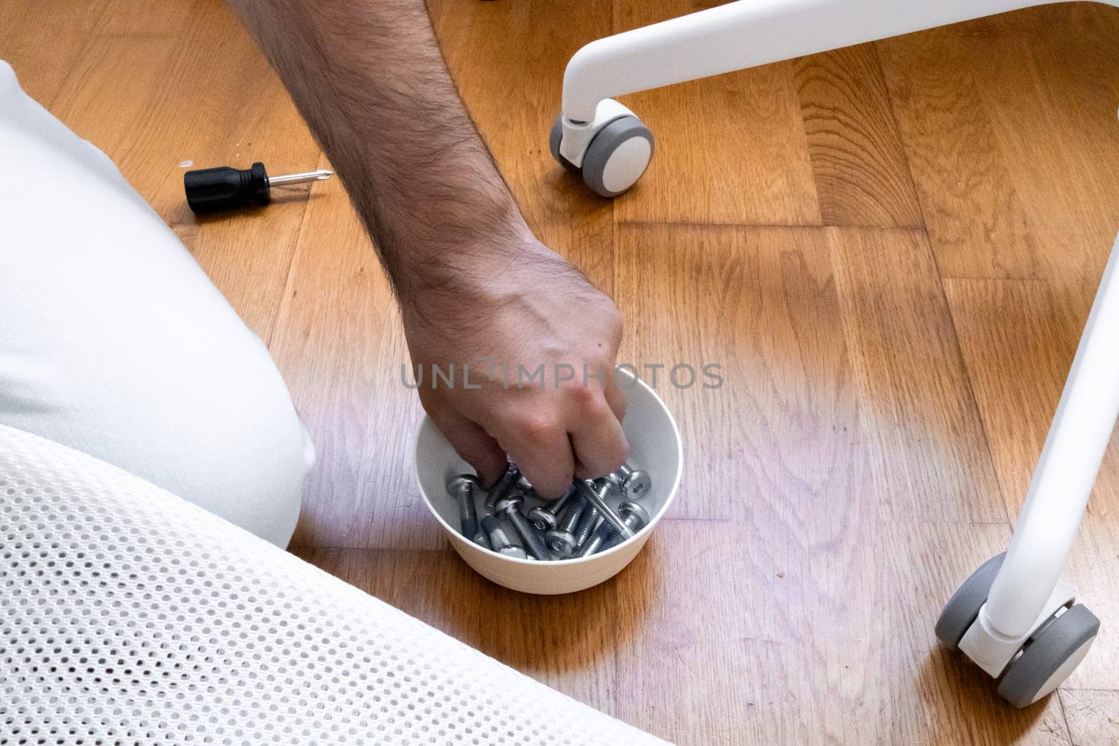 Adult male assembling furniture. Hand grabbing screws on wooden floor near white chair wheel.