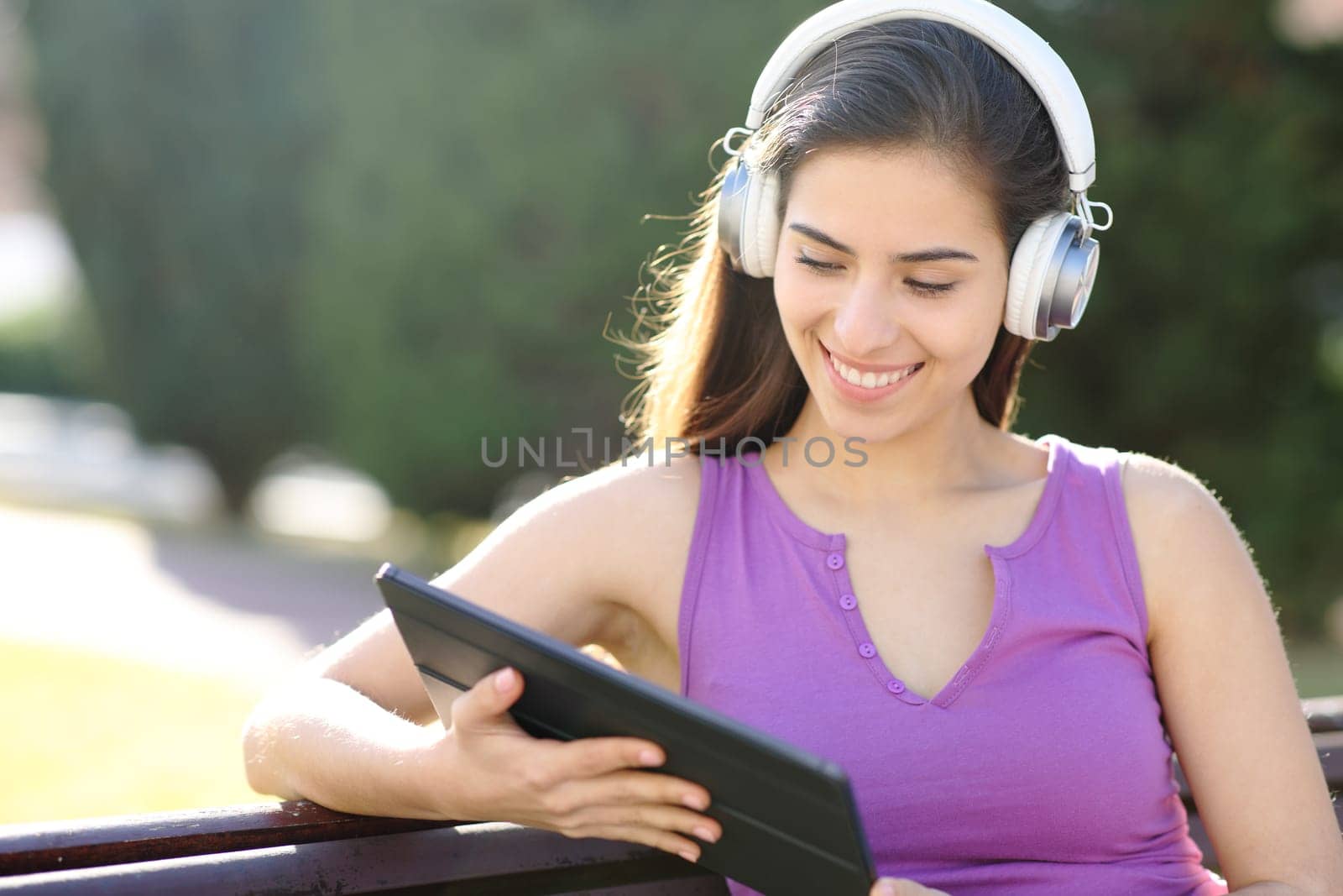 Happy woman watching media on tablet with headphone sitting in a park