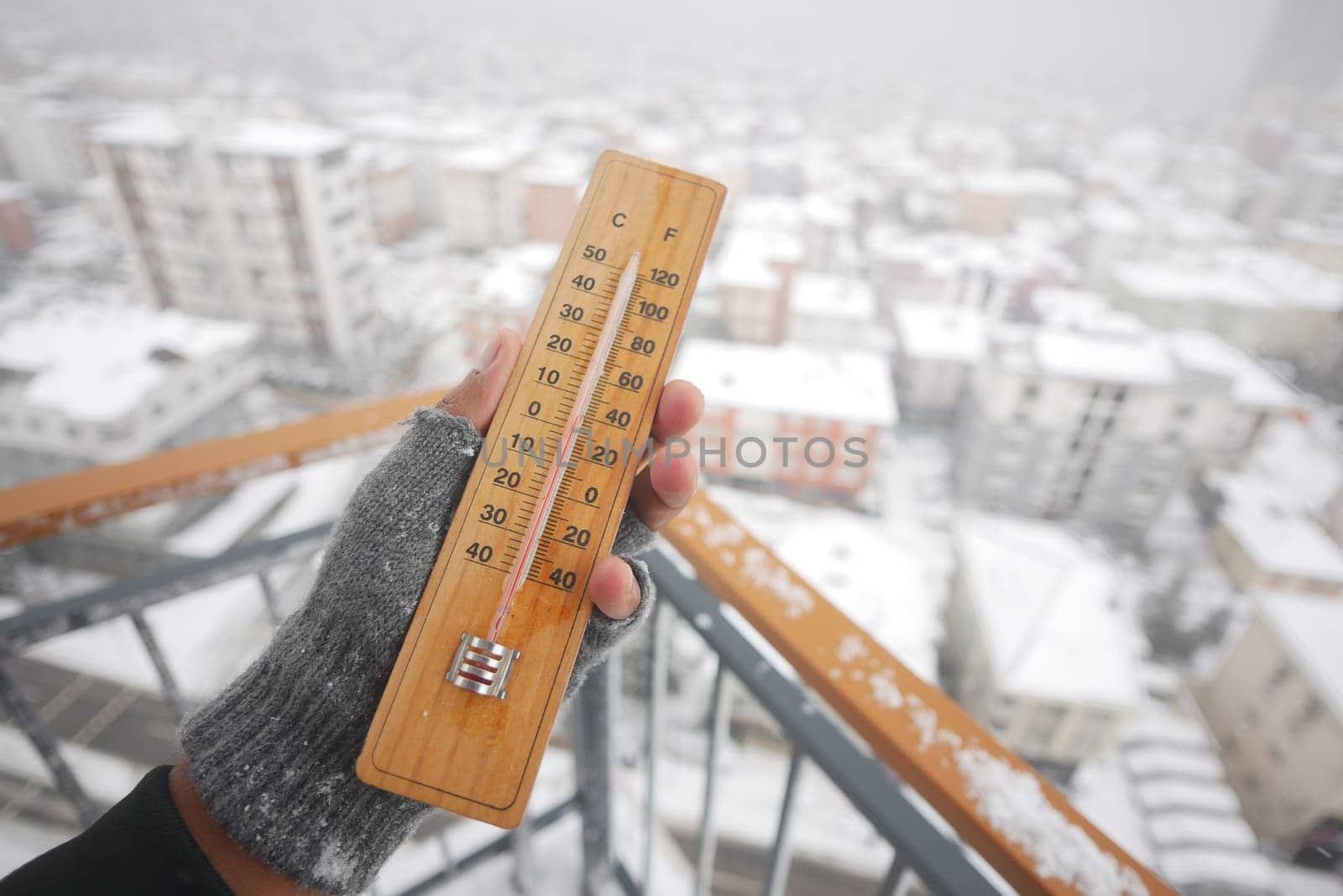 A person holds a thermometer as snow blankets the cityscape below.