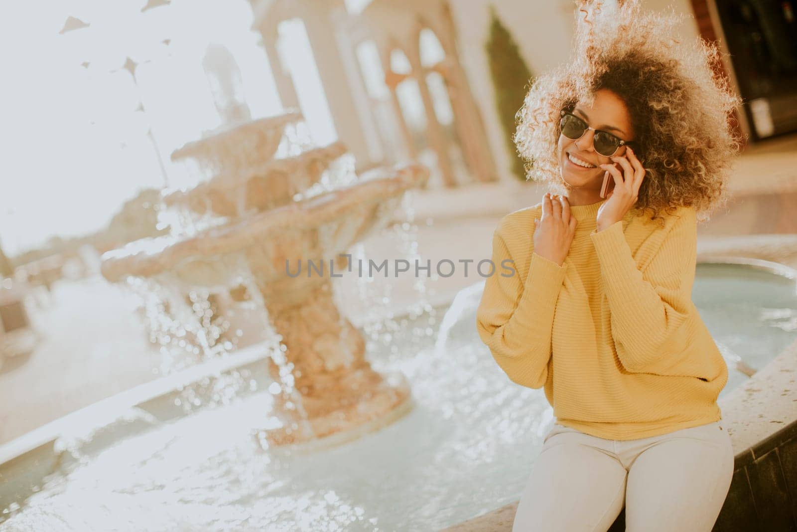Aftican american young woman sits near fountain and talks on smartphone
