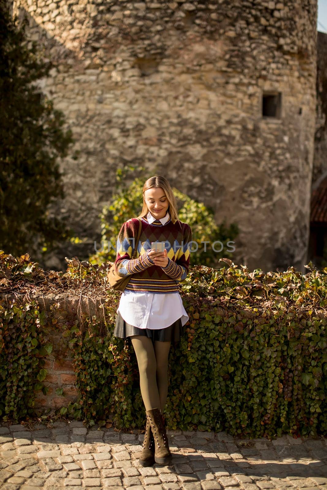 Pretty young woman standing with mobile phone on street at sunny autumn day
