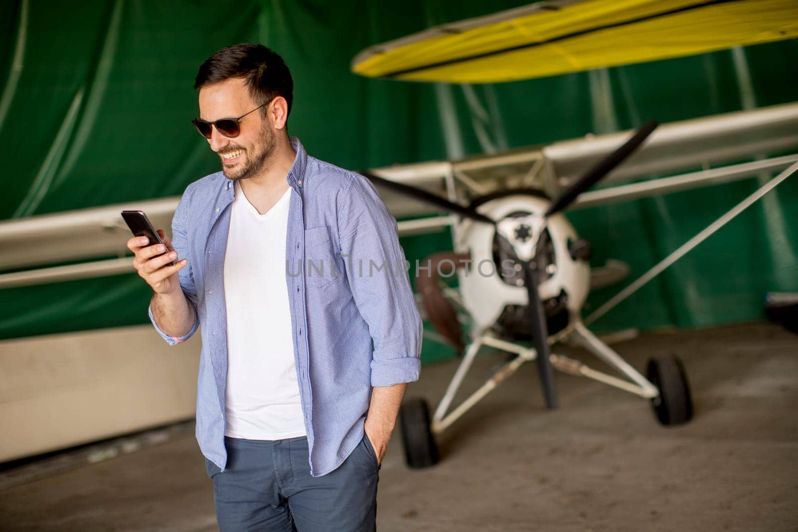 Young handsome young pilot checking airplane in the hangar and using mobile phone