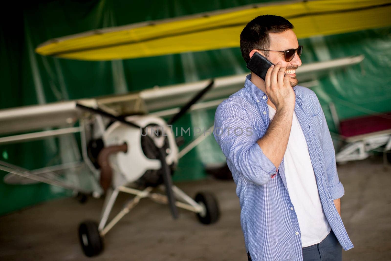 Young handsome young pilot checking airplane in the hangar and using mobile phone