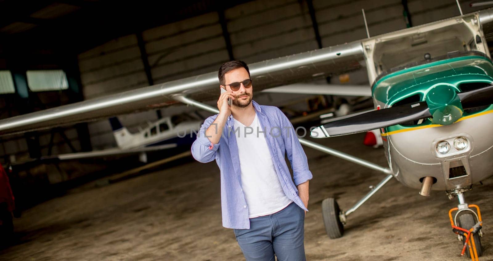 Young handsome young pilot checking airplane in the hangar and using mobile phone