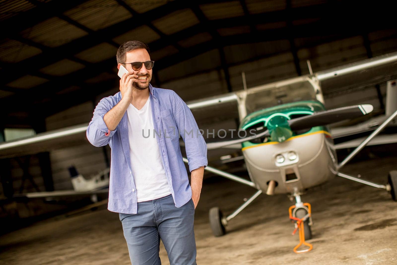 Young handsome young pilot checking airplane in the hangar and using mobile phone