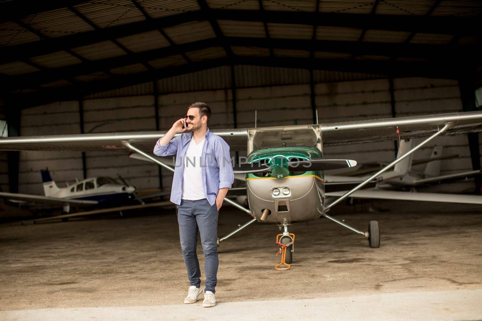 Young handsome young pilot checking airplane in the hangar and using mobile phone