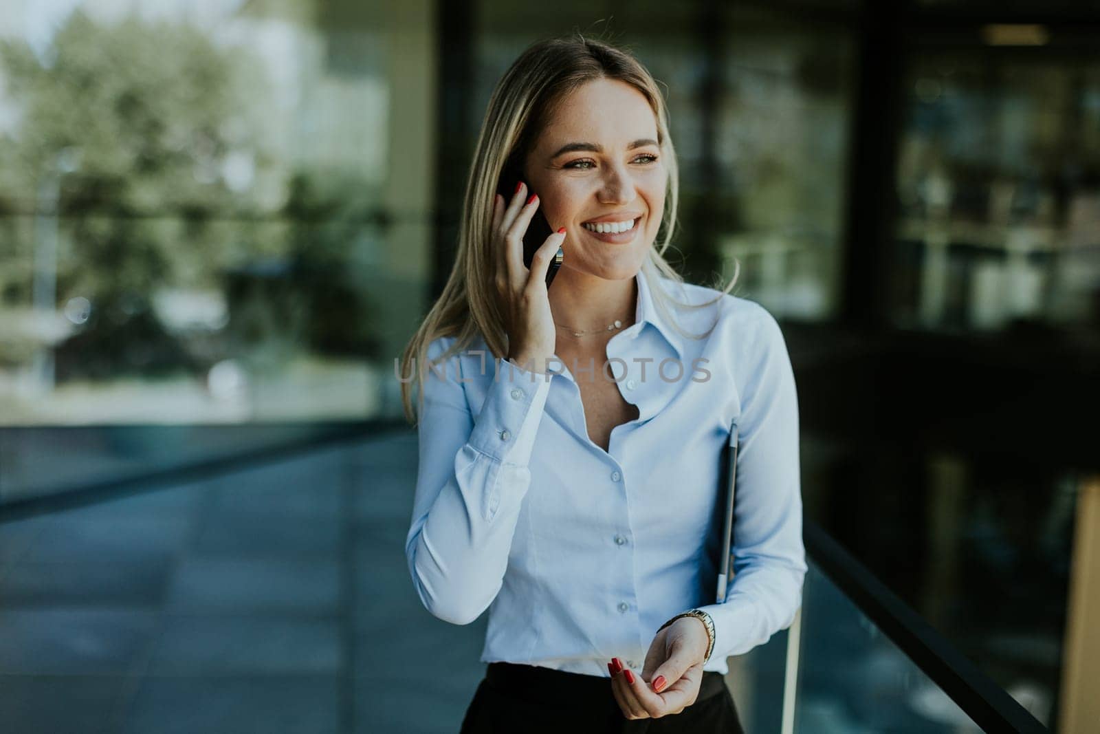 A young businesswoman smiles confidently while talking on her phone inside a sleek, modern office building, with cityscape reflections in the glass.