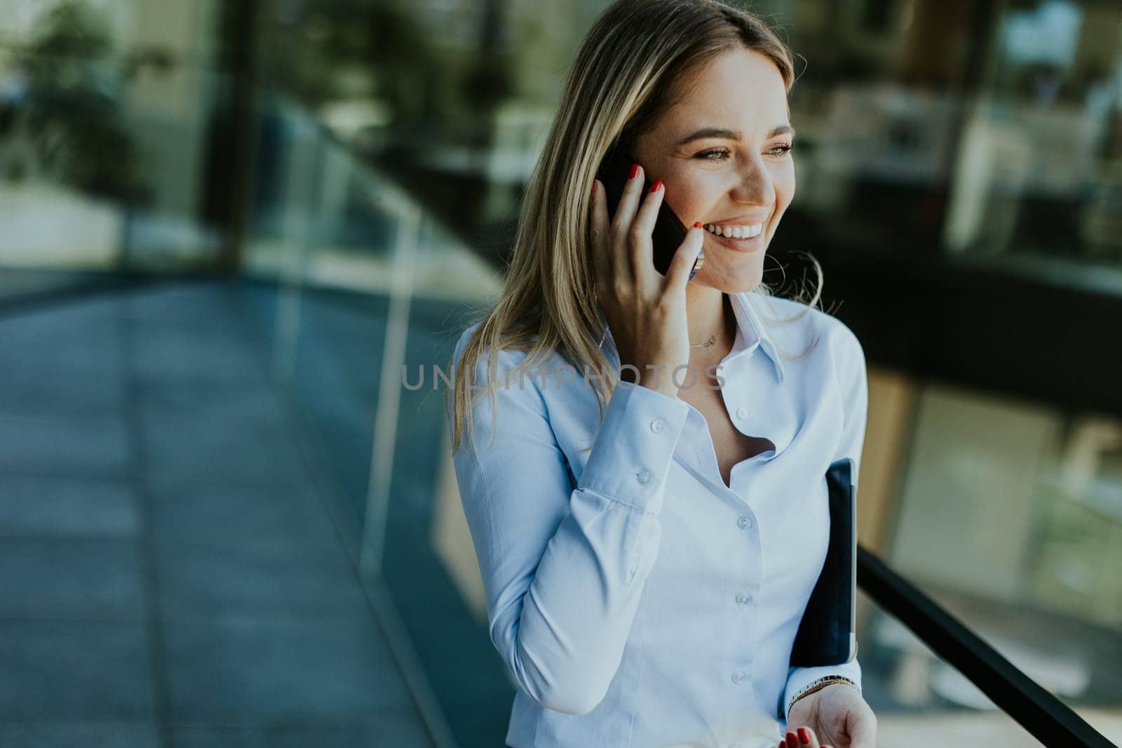 A young businesswoman smiles confidently while talking on her phone inside a sleek, modern office building, with cityscape reflections in the glass.