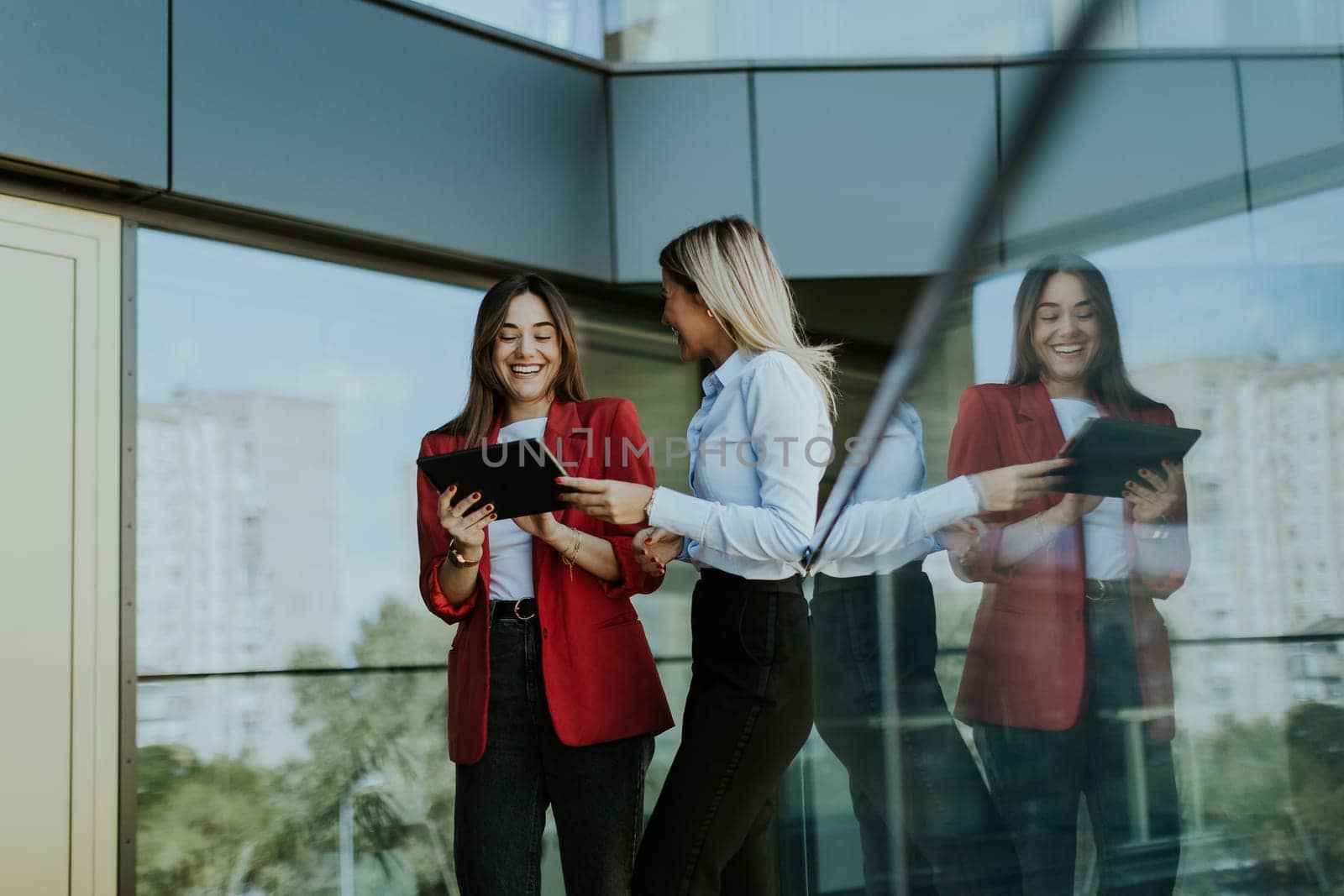 Two professional women stand outside an office building, engaging in a lively discussion while using digital tablets. Their reflections are visible on the glass facade