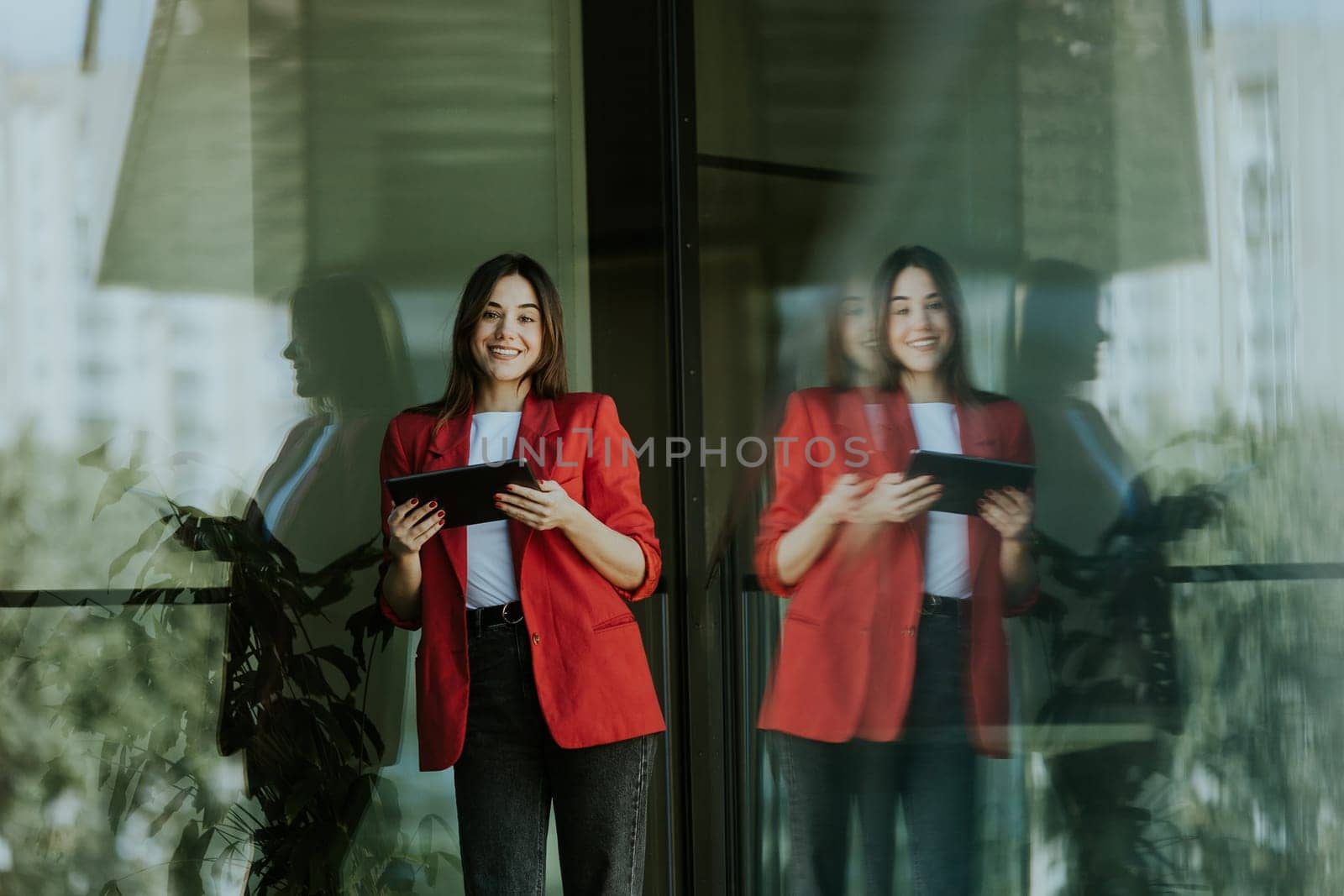 A smiling professional woman stands near a modern glass office building, holding a tablet and looking confident during the evening.