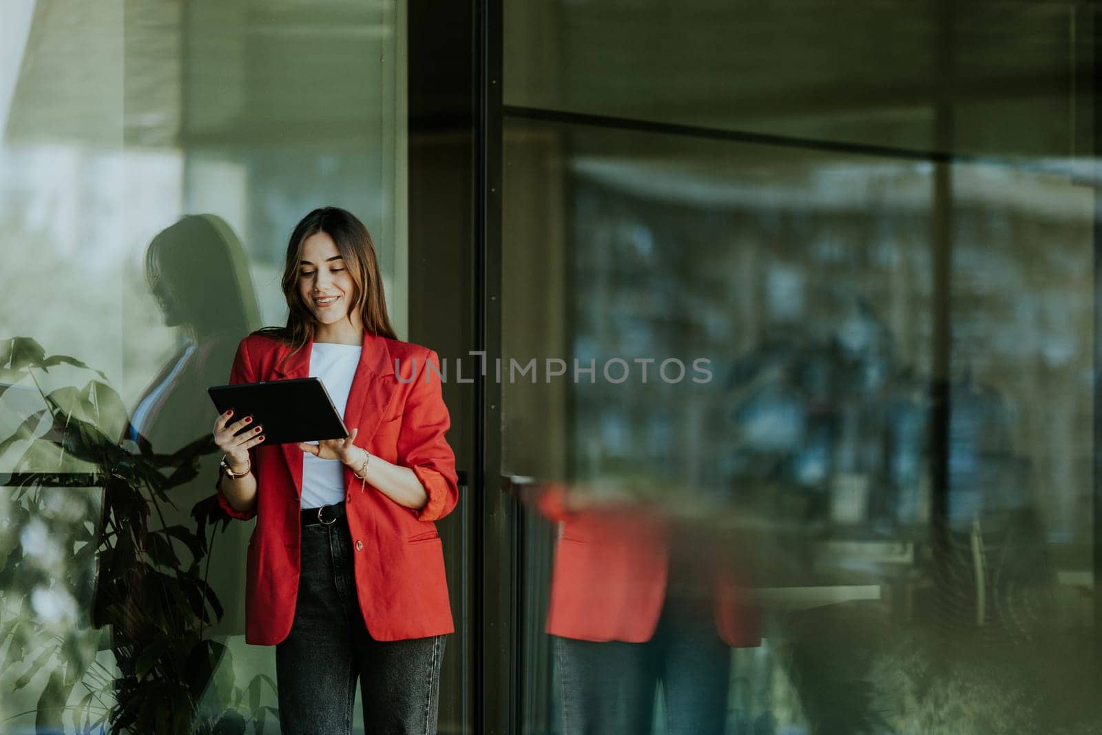 A smiling professional woman stands near a modern glass office building, holding a tablet and looking confident during the evening.