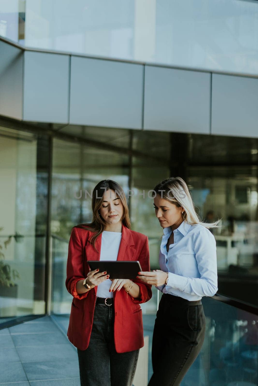 Two professional women stand outside an office building, engaging in a lively discussion while using digital tablets. Their reflections are visible on the glass facade